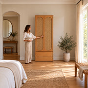 Alvarario 2 Door Wooden Cane Wardrobe with Drawer with a woman standing beside the cabinet, showing a natural wood finish.