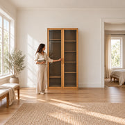 Porteriy 2 Door Wooden Closet Cabinet beside a woman reaching toward the door, featuring two smoke-grey glass doors.